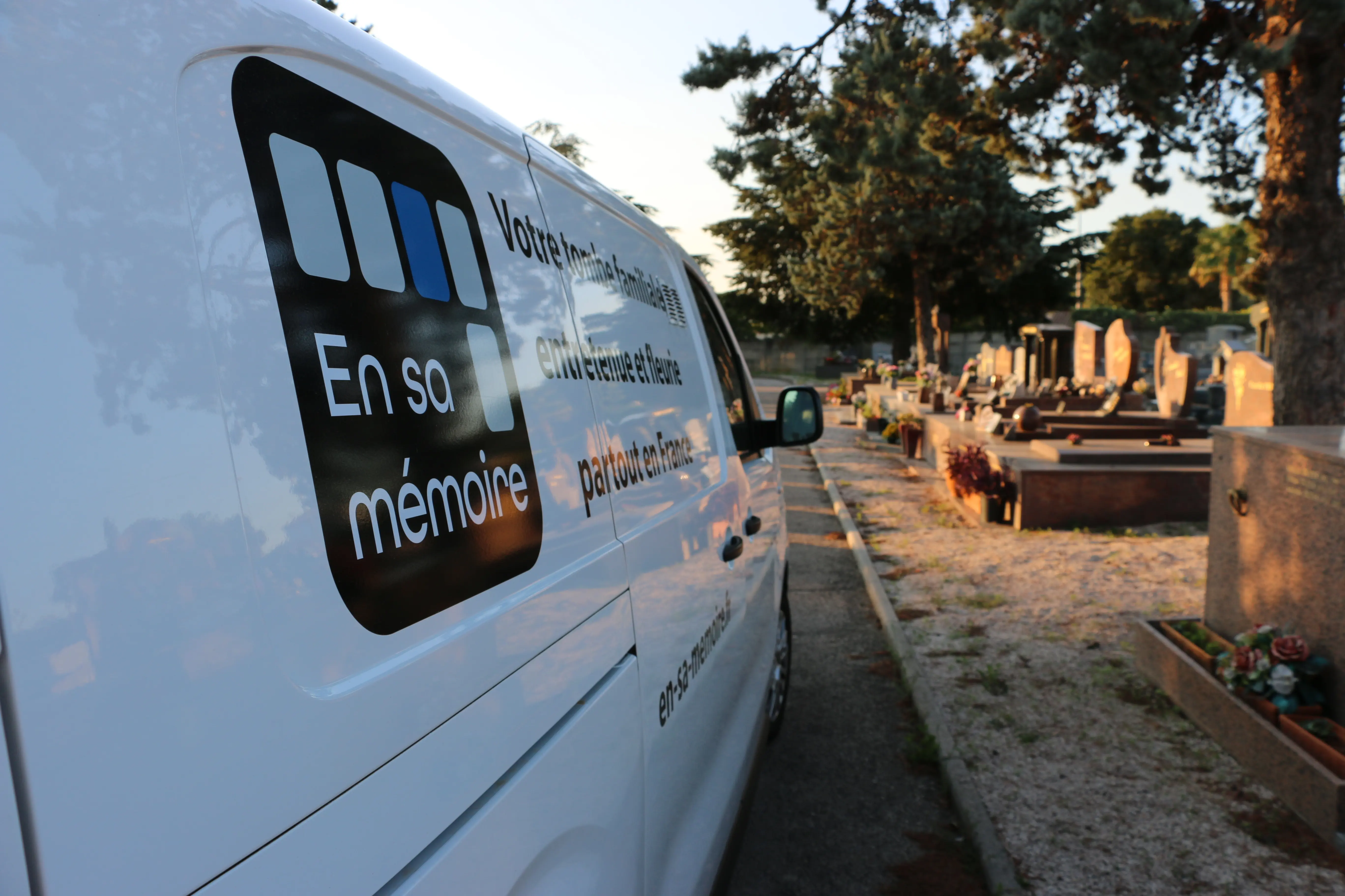 Van blanche avec logo "En sa mémoire" sur le côté, véhicule d’entretien près de tombes et d’urnes, prêt pour le fleurissement.
