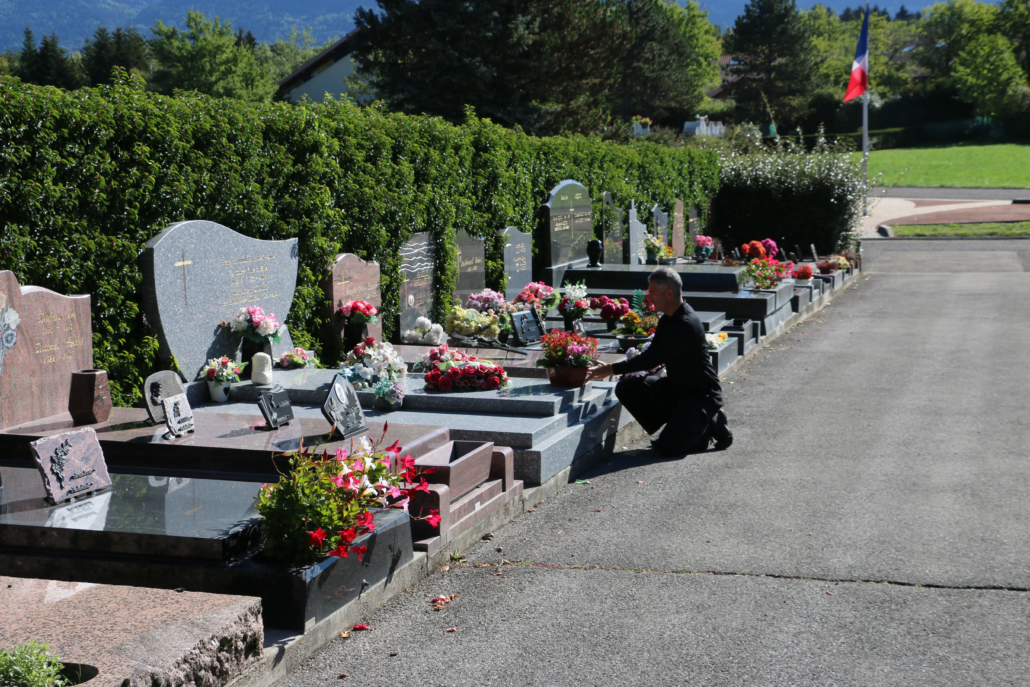 Homme agenouillé près des tombes alignées dans un cimetière, fleurs et bouquets décorent les plaques tombales, drapeau au loin. Texte lié : comment entretenir une tombe.