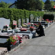 Homme agenouillé près des tombes alignées dans un cimetière, fleurs et bouquets décorent les plaques tombales, drapeau au loin. Texte lié : comment entretenir une tombe.