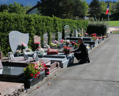Homme agenouillé près des tombes alignées dans un cimetière, fleurs et bouquets décorent les plaques tombales, drapeau au loin. Texte lié : comment entretenir une tombe.