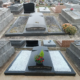 Tombe rectangulaire en pierre sombre dans un cimetière, entourée de gravats et d'autres monuments. Déco fleurie au centre avec un petit arrangement végétal coloré, et un lit de gravier blanc sur les côtés.