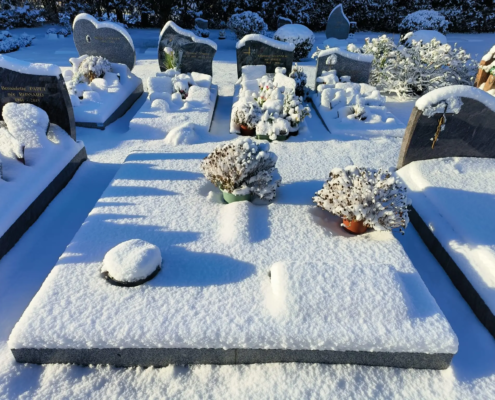 Tombeau enneigé dans un cimetière, avec des pierres tombales à l'arrière et des pots de fleurs blanches couvertes de neige.