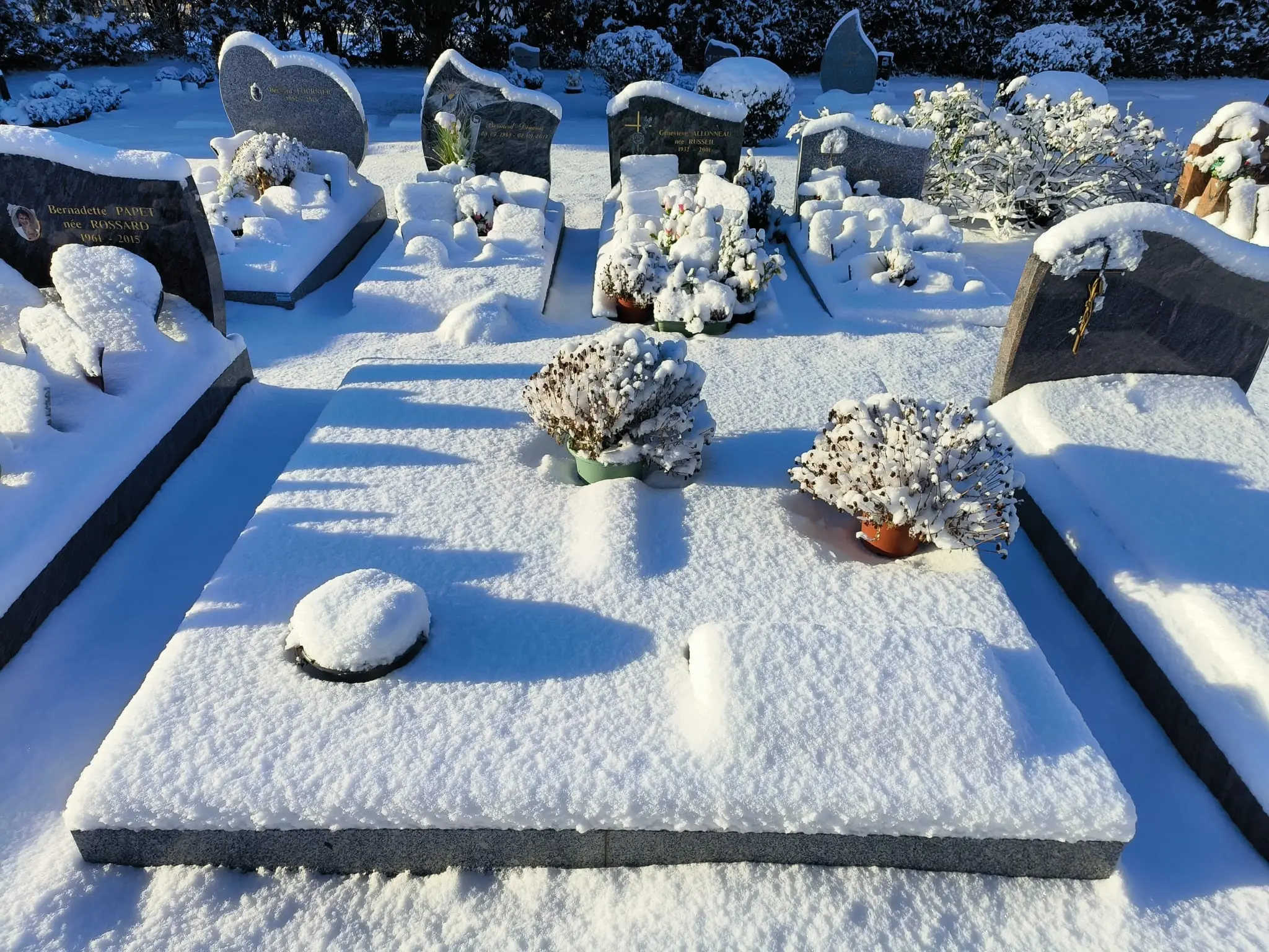 Tombeau enneigé dans un cimetière, avec des pierres tombales à l'arrière et des pots de fleurs blanches couvertes de neige.