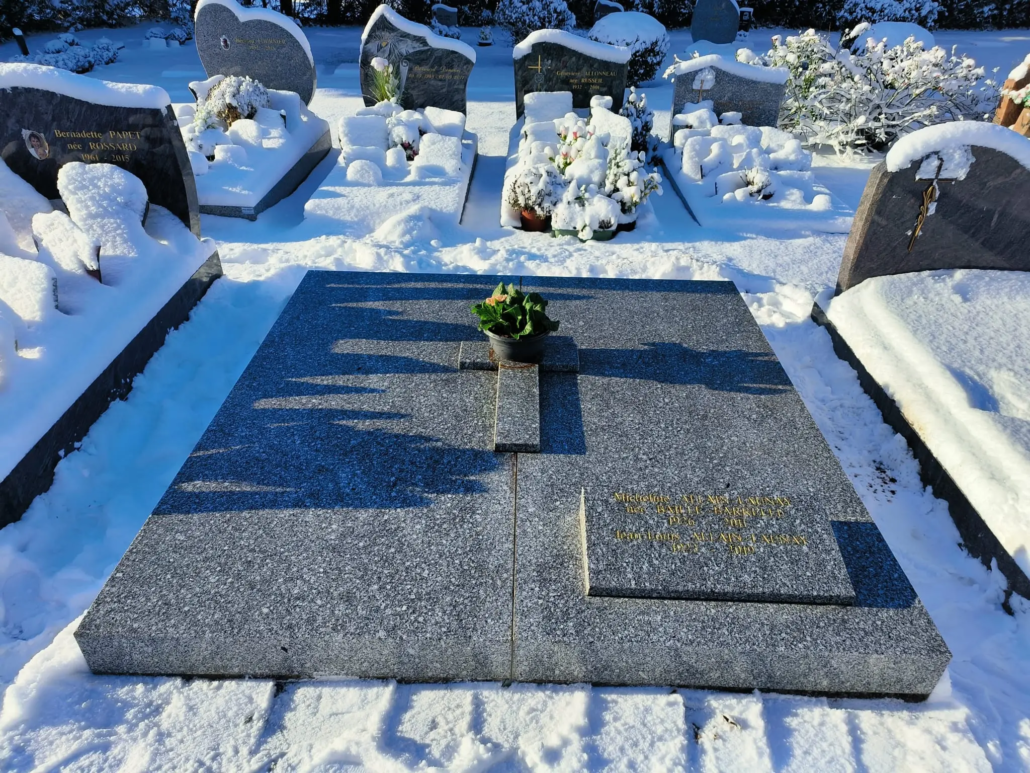 Tombe en granit décorée d'une petite plante en pot au centre, entourée de tombes enneigées et de vases fleuris, ambiance hivernale. Fleur pour le cimetière en hiver intégrée naturellement.