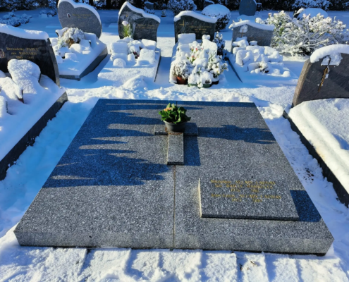 Tombe en granit décorée d'une petite plante en pot au centre, entourée de tombes enneigées et de vases fleuris, ambiance hivernale. Fleur pour le cimetière en hiver intégrée naturellement.