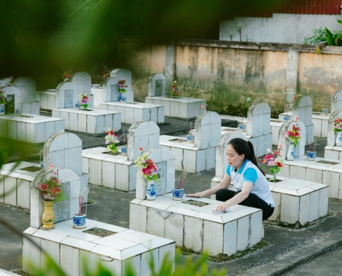 Travailleuse nettoyant et fleuri des tombes dans un cimetière, rangées de monuments funéraires blancs et vases de fleurs colorées.