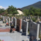 Entrée d’un cimetière bien entretenu avec des tombes en pierre, fleurs et verdure, sous un ciel bleu et des montagnes en arrière-plan.