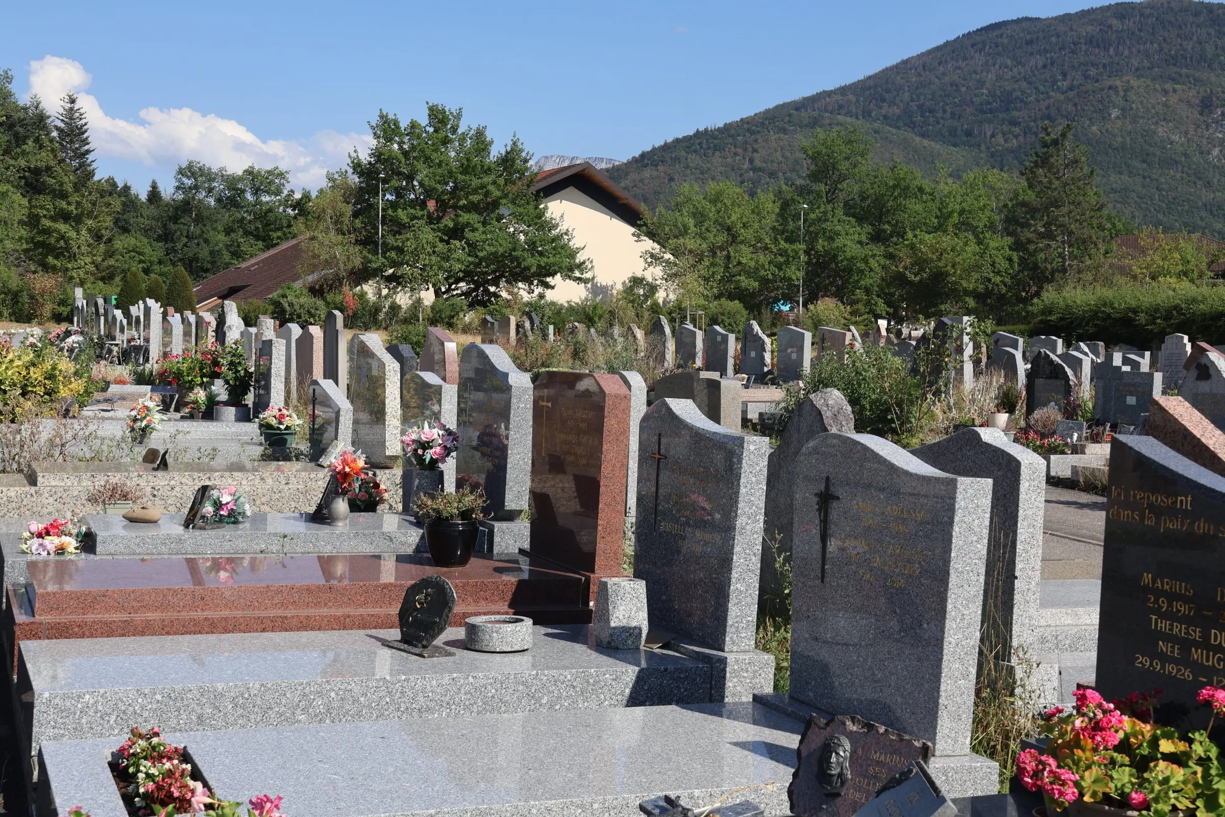 Entrée d’un cimetière bien entretenu avec des tombes en pierre, fleurs et verdure, sous un ciel bleu et des montagnes en arrière-plan. Entrée d’un cimetière bien entretenu avec des tombes en pierre, fleurs et verdure, sous un ciel bleu et des montagnes en arrière-plan.