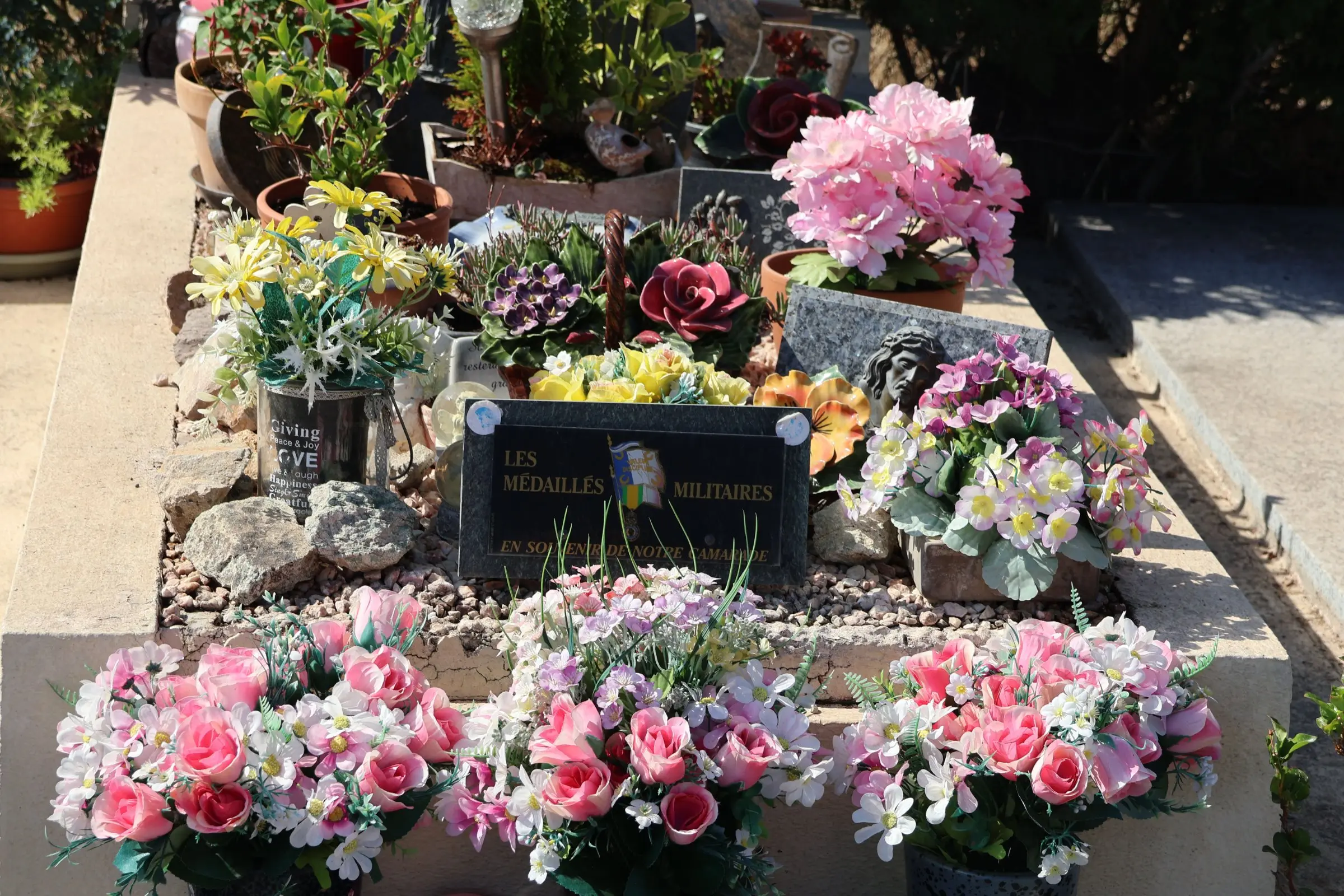 Tombe décorée avec des arrangements floraux variés : roses, fleurs pastel et plantes succulentes disposées autour d’une stèle gravée. Tombe décorée avec des arrangements floraux variés : roses, fleurs pastel et plantes succulentes disposées autour d’une stèle gravée.