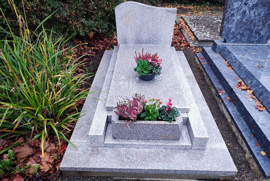 Tombe en pierre grise avec des fleurs plantées dans des pots au sommet et sur le rebord, entourée de feuilles mortes et d’un