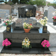 Tombe en pierre noire avec motifs gravés et plaques dorées, entourée de vases et bouquets colorés sur le devant. nettoyage de tombe tarif intégré.