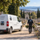 Voiture utilitaire blanche avec logo "En sa mémoire" garée dans un cimetière fleuri, un salarié porte des pots de fleurs.