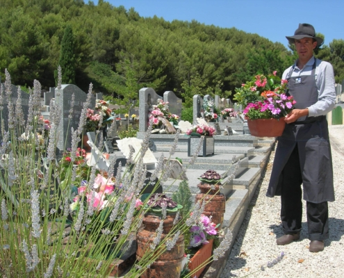 Un homme portant un tablier et un chapeau dépose un pot de fleurs sur une tombe au cimetière.