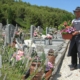 Un homme portant un tablier et un chapeau dépose un pot de fleurs sur une tombe au cimetière.