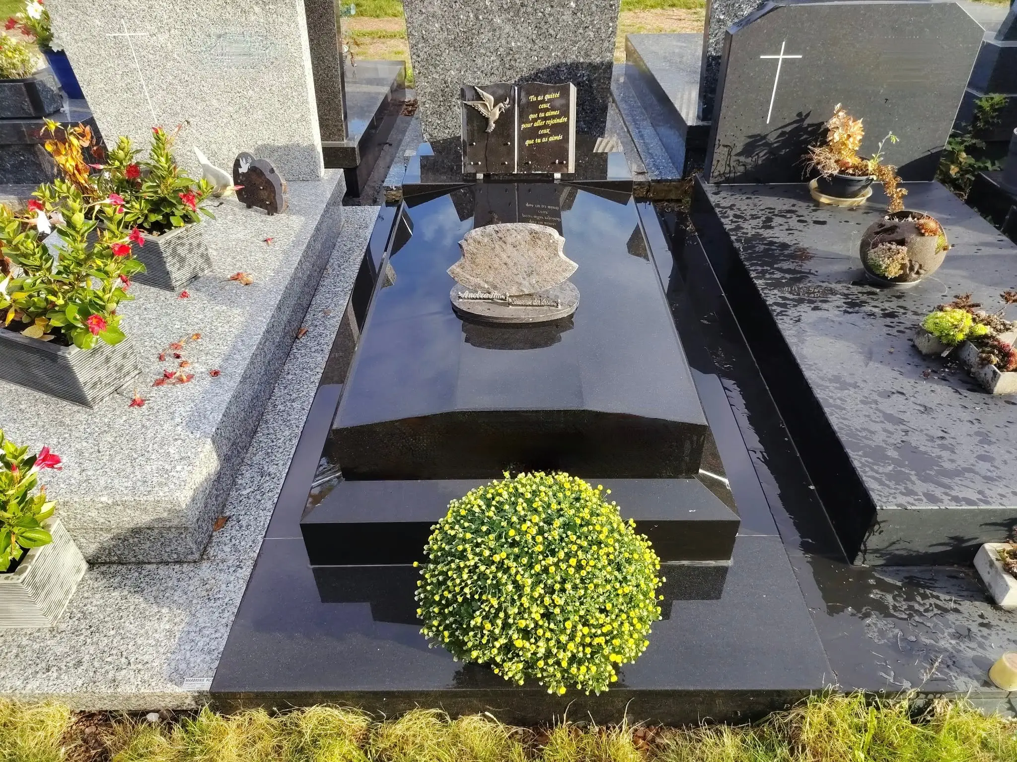Tombe familiale en pierre noire polie avec entourage et végétation décorative, fleurs artificielles et verdure décorative Tombe familiale en pierre noire polie avec entourage et végétation décorative, fleurs artificielles et verdure décorative