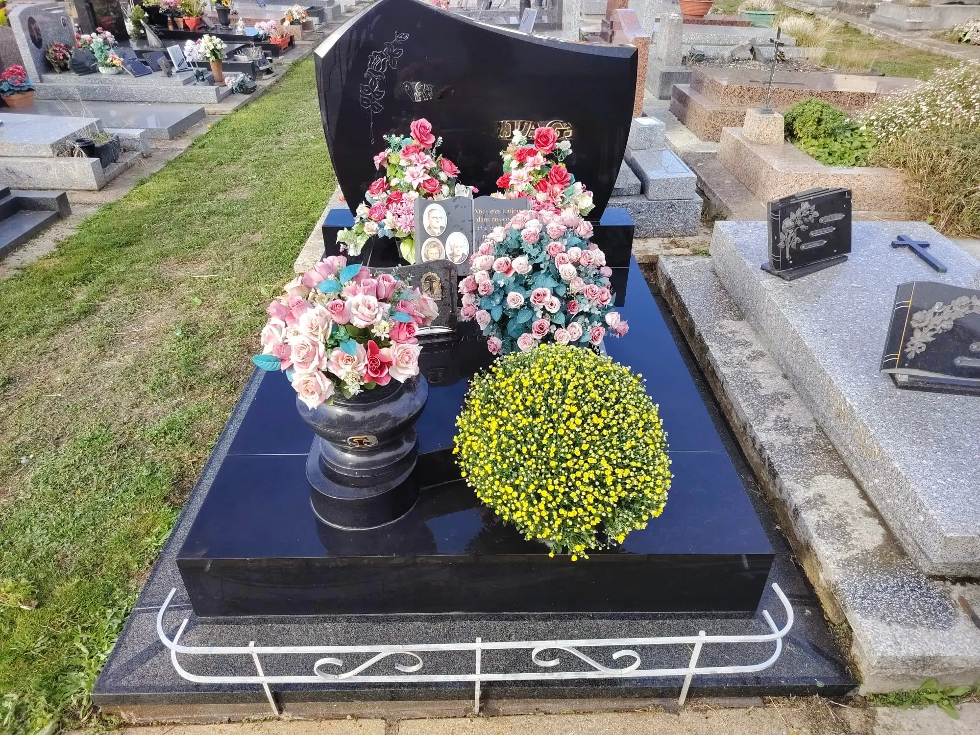 Tombe avec tête gravée, vase rond de fleurs roses et bouquets sur un socle noir poli; bouquet jaune ronds et clôture blanche Tombe avec tête gravée, vase rond de fleurs roses et bouquets sur un socle noir poli; bouquet jaune ronds et clôture blanche